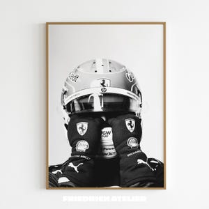 May include: Black and white framed print of a racing driver. The driver is wearing a helmet and gloves with sponsor logos. The helmet has a visor and the gloves cover the driver's face. The print is in a brown frame.