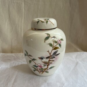 Könnte beinhalten: Eine cremefarbene Keramikdose mit Deckel, verziert mit einem gemalten Design eines Vogels auf einem blühenden Zweig. Die Dose hat einen goldenen Rand und eine glänzende Oberfläche. Der Vogel hat graue, braune und weiße Federn, mit rosa Blüten.