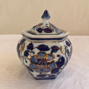 Könnte beinhalten: Eine weiß-blaue Keramikdose mit Deckel. Die Dose hat ein Blumendesign mit goldenen Akzenten. Der Deckel hat eine spitze Spitze. Die Dose dient wahrscheinlich zur Aufbewahrung oder Dekoration.
