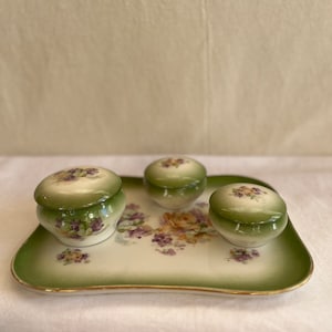 Könnte beinhalten: Ein antikes Porzellan-Schmucktablett mit drei kleinen, mit Deckel versehenen Dosen. Das Tablett und die Dosen sind mit einem floralen Muster in Grün-, Lila- und Gelbtönen verziert.