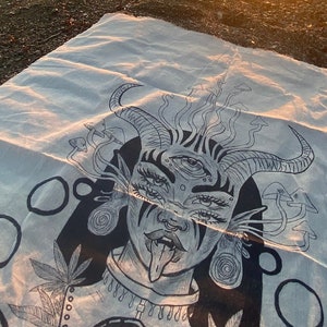 Puede incluir: Una toalla de playa blanca con una ilustraci&oacute;n en blanco y negro de una mujer con cuernos, m&uacute;ltiples ojos y la lengua fuera. La ilustraci&oacute;n est&aacute; rodeada de c&iacute;rculos y hojas.