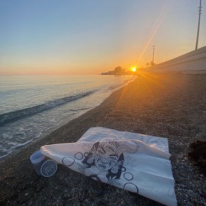 Puede incluir: Una bolsa de lona blanca con una ilustraci&oacute;n en blanco y negro de una cara con tres ojos y cuernos. La bolsa est&aacute; tumbada en una playa con una taza y la palabra "folk" impresa en ella.