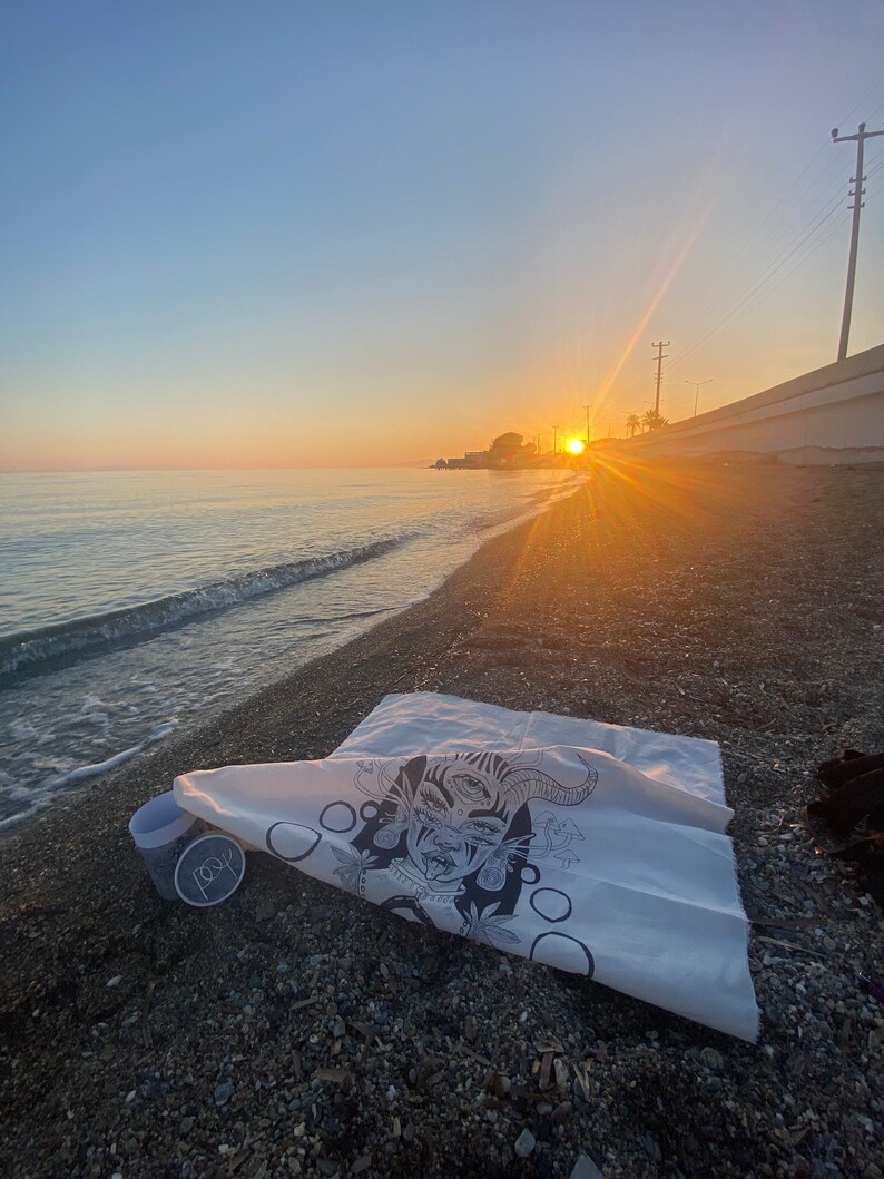 Puede incluir: Una bolsa de lona blanca con una ilustraci&oacute;n en blanco y negro de una cara con tres ojos y cuernos. La bolsa est&aacute; tumbada en una playa con una taza y la palabra "folk" impresa en ella.