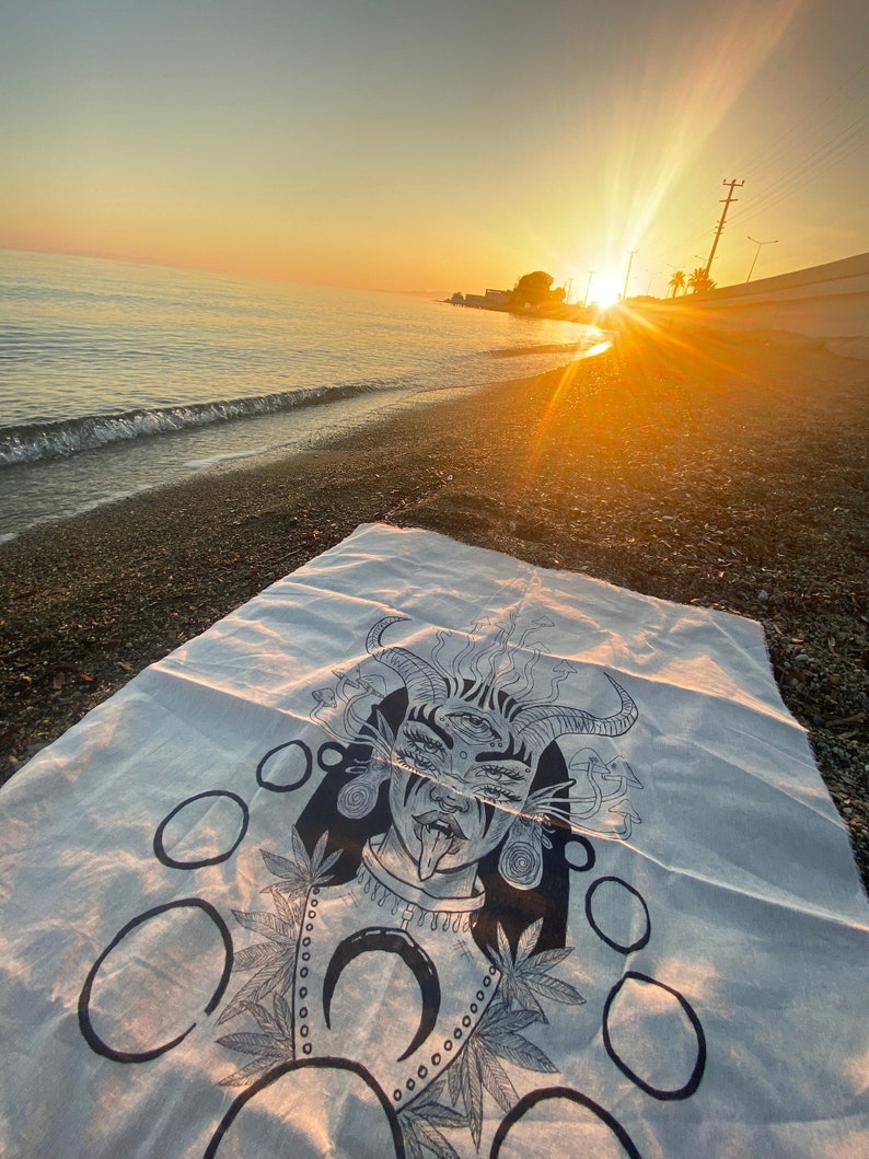 Puede incluir: Una toalla de playa blanca con una ilustraci&oacute;n en blanco y negro de una mujer con cuernos, m&uacute;ltiples ojos y la lengua fuera. La ilustraci&oacute;n est&aacute; rodeada de c&iacute;rculos y hojas.