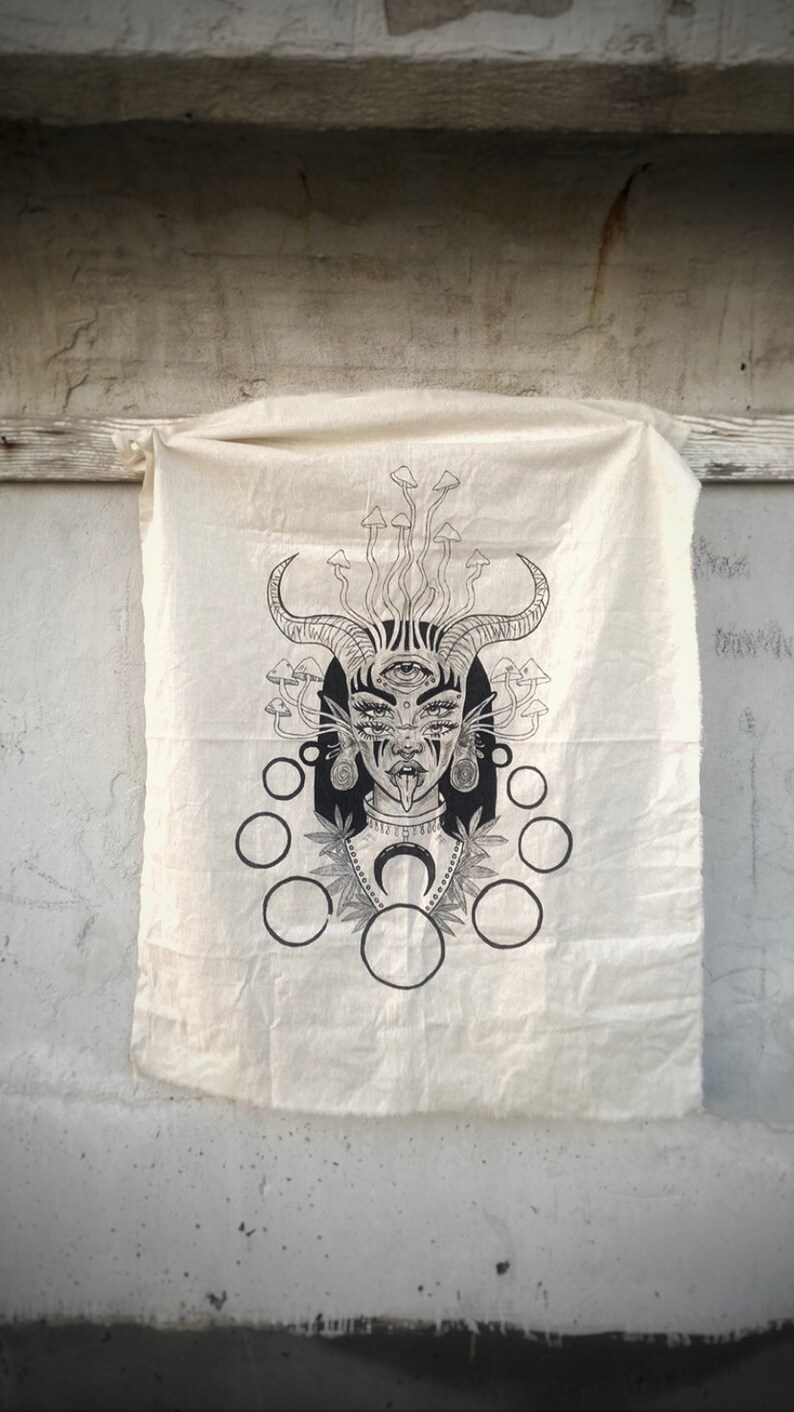 Puede incluir: Una ilustraci&oacute;n en blanco y negro de una mujer con cuernos, un tercer ojo y la lengua fuera. Est&aacute; rodeada de setas y c&iacute;rculos. La ilustraci&oacute;n est&aacute; impresa en un lienzo blanco.