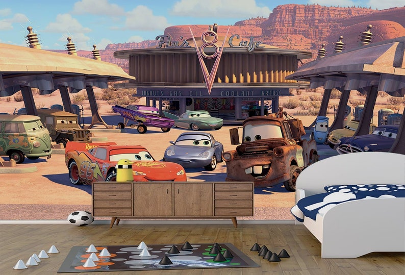 K&ouml;nnte beinhalten: Ein farbenfrohes Wandbild zum Thema Cars mit verschiedenen Cartoon-Autos, darunter Mater, Lightning McQueen und ein blaues Auto mit rotem Streifen. Das Wandbild ist vor dem Hintergrund einer W&uuml;stenlandschaft mit einer Tankstelle und einem Schild mit der Aufschrift "Flo's V8 Cafe" platziert.