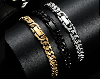 May include: Three chain link bracelets in gold, black, and silver are displayed on a black surface. The gold bracelet has a gold clasp, the black bracelet has a black clasp, and the silver bracelet has a silver clasp. A wristwatch is visible in the background.