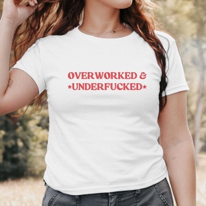 Op de afbeelding: Witte baby-T-shirt met de tekst "OVERWORKED & UNDERFUCKED" in het rood. De tekst staat in het midden op de voorkant van de shirt. Het shirt heeft een ronde hals en korte mouwen. Het model draagt een grijze broek.