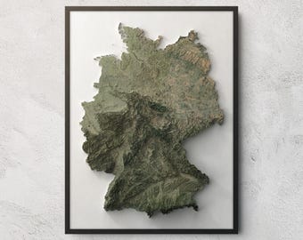 May include: A framed wall art piece featuring a three-dimensional topographic relief map of Germany. The map displays detailed terrain textures in shades of green, brown, and grey, set against a plain white background within a thin black frame.