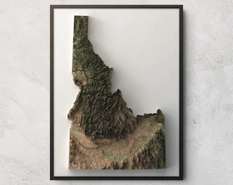 May include: A framed three-dimensional relief map of the state of Idaho. The map features detailed topographical textures with dark green forested mountain ranges in the north and tan, arid desert terrain in the southern regions of the state.