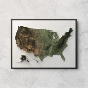 May include: A framed wall art piece featuring a three-dimensional relief map of the United States, including Alaska and Hawaii. The map displays varied terrain textures and colors, transitioning from brown arid regions to green forested areas.