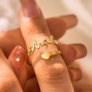 Può includere: Un anello color oro con il nome "Angela" in corsivo. L'anello presenta un dettaglio a forma di cuore e viene indossato su un dito. È un gioiello personalizzato.