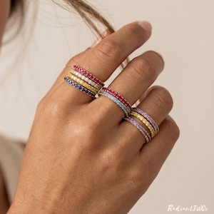 Könnte beinhalten: Eine Hand präsentiert mehrere farbenfrohe Edelsteinringe. Die Ringe weisen eine Vielzahl von Farben auf, darunter Rot, Blau, Gelb und Lila. Die Ringe sind in Gold gefasst. Das Bild ist gut beleuchtet und zeigt das Funkeln der Edelsteine.