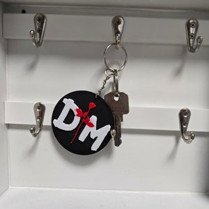Könnte beinhalten: Ein schwarz-weißer Schlüsselanhänger mit den Buchstaben "DM" und einem roten Blumendesign. Der Schlüsselanhänger hängt an einem silbernen Schlüsselring mit einem Schlüssel. Der Schlüsselanhänger befindet sich in einem weißen Schlüsselhalter mit silbernen Haken.