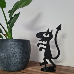 Peut inclure: Sculpture en métal noir d'un diable de dessin animé avec une longue queue et une flèche pointue à l'extrémité. Le diable a un seul œil et se tient sur une base ronde. Une plante en pot est en arrière-plan.
