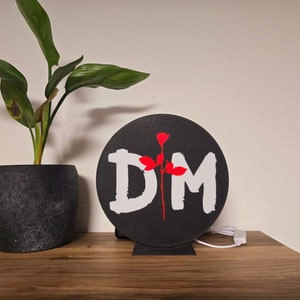 Könnte beinhalten: Eine schwarze runde Lampe mit den Buchstaben "DM" in Weiß, einer roten Rose und einem Ständer. Die Lampe steht auf einer Holzoberfläche neben einer Topfpflanze mit grünen Blättern. Ein weißes Kabel geht von der Lampe aus.