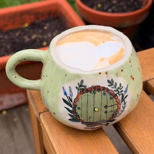 Peut inclure: Une tasse en céramique vert clair avec une forme ronde et une anse. La tasse présente un dessin peint à la main d'une porte verte avec une couronne florale. La tasse est remplie d'un cappuccino et est posée sur une surface en bois.