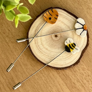 Può includere: Tre spille decorative con un design di ape, fiore e favo. Le spille sono argentate con un'ape nera e bianca, un fiore bianco con un centro giallo e un favo marrone e giallo.