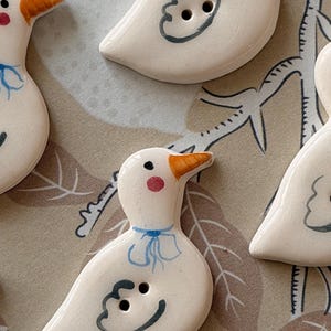 Peut inclure: Plusieurs boutons en céramique en forme de canard. Chaque bouton est blanc avec un bec orange, un nœud bleu et une joue rose. Les boutons ont deux trous et un contour gris peint. Le fond est un tissu à motifs.