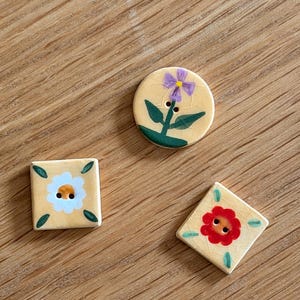 Puede incluir: Tres botones de cerámica con base color crema y acabado de esmalte craquelado. Un botón redondo presenta una flor morada, un botón cuadrado muestra una flor blanca y un botón en forma de rombo tiene una flor roja, todos con hojas verdes.