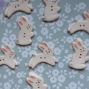 May include: A collection of off-white ceramic bunny-shaped buttons. Each button features pink-painted ears, black dot eyes, and a small black tail. The buttons are arranged on a light blue floral patterned surface.