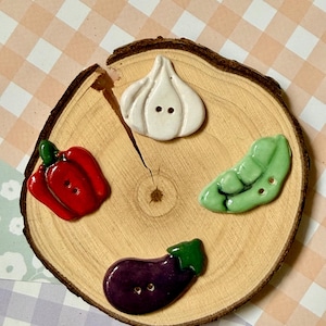Pode incluir: Quatro botões de cerâmica em forma de vegetais, incluindo um bulbo de alho branco, um pimentão vermelho, uma vagem de ervilha verde e uma berinjela roxa. Cada botão possui dois pequenos orifícios para costura e um acabamento liso e brilhante.