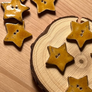 Könnte beinhalten: Nahaufnahme von sternförmigen Knöpfen mit rustikalem, glasiertem Finish. Die Knöpfe haben eine warme gelb-orange Farbe und zwei Löcher zum Annähen. Einige Knöpfe liegen auf einer Holzoberfläche, andere sind auf einer Holzscheibe angeordnet.