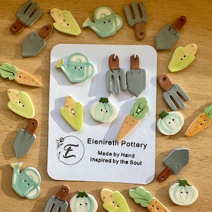 May include: A collection of hand-painted ceramic buttons featuring garden-themed designs, including carrots, onions, watering cans, and gardening tools. The buttons are arranged in a circle around a card with the text "Elenireth Pottery, Made by Hand, Inspired by the Soul."
