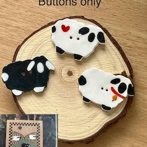 Peut inclure: Trois boutons en forme de mouton, noirs et blancs, avec un cœur rouge et des taches noires. Les boutons sont sur une tranche de bois. Le texte "Buttons only" est en haut. Un motif de point de croix est en bas à gauche.