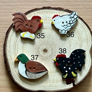 May include: Four wooden buttons shaped like farm animals: a rooster, a hen, and a duck. The rooster is brown with a red comb, the hen is white with black detailing, the duck is brown and white, and the other rooster is black with white dots. Numbers 35, 36, 37, and 38 are printed on the wood.