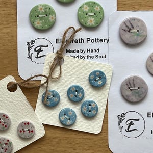 May include: Assortment of handmade ceramic buttons with floral and botanical designs. The buttons are displayed on white cards with the text "Elfreth Pottery, Made by Hand, Inspired by the Soul".