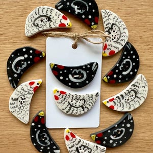 Op de afbeelding: Een set van tien keramische knoppen in de vorm van halve manen, elk met een handgeschilderd ontwerp van een kip. De knoppen zijn in twee kleurschema's: zwart met witte stippen en wit met zwarte en grijze markeringen. Elke knop heeft een rode kam en een gele snavel.
