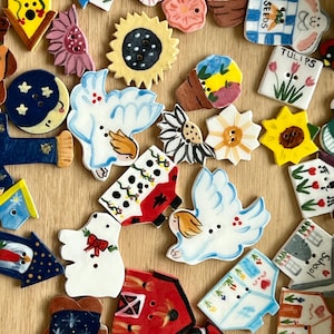 May include: A selection of hand-painted ceramic buttons in diverse shapes, including flowers, houses, and birds. The buttons display intricate details and a range of colours, such as yellow, blue, red, and white. Some buttons have text.