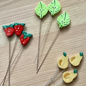 May include: A set of nine decorative pins featuring small, hand-painted shapes. Three pins are red strawberries with green tops, three are light green leaves with smiling faces, and three are yellow pears with green stems, all attached to long metal needles.