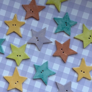 May include: A collection of star-shaped buttons in various colors including yellow, teal, orange, and light purple. Each button features two small holes in the center for attachment. The buttons are arranged in a scattered pattern across the frame.