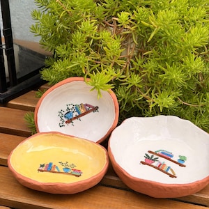 May include: Three small, hand-formed terracotta bowls with irregular, wavy edges. Two bowls have white interiors, and one has a yellow interior. Each bowl features a hand-painted illustration of wooden shelves holding colorful books and potted plants.