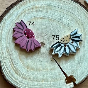 Op de afbeelding: Twee decoratieve bloemvormige knopen. Eén is roze met een gestippeld midden en het nummer 74. De andere is wit en grijs met een gestippeld midden en het nummer 75. Beide liggen op een houten schijf.