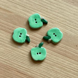 Peut inclure: Quatre boutons verts en forme de pomme avec des tiges vert foncé et de petits accents blancs. Chaque bouton a deux trous pour la couture. Les boutons sont disposés sur une surface en bois brun clair.