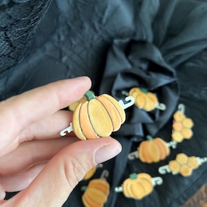 Könnte beinhalten: Eine Sammlung von orangefarbenen und gelben Kürbis-Anstecknadeln mit grünen Stielen. Die Anstecknadeln haben eine silberfarbene Rückseite. Die Anstecknadeln befinden sich auf einem schwarzen Hintergrund. Die Anstecknadeln sind wahrscheinlich für Halloween- oder Herbst-Bastelarbeiten.