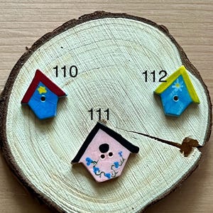 May include: Three miniature, hand-painted birdhouses with varying colors and designs are displayed on a wooden slice. The birdhouses have red, yellow, and black roofs, with blue and pink bodies. Each birdhouse has a unique number: 110, 111, and 112.