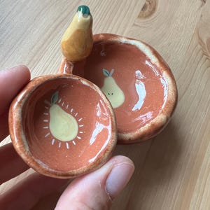 Könnte beinhalten: Handgefertigte Keramik-Schmuckschalen mit Birnen-Motiv. Die Schalen sind terrakottafarben mit einem Birnen-Design in der Mitte. Eine Schale hat einen birnenförmigen Griff. Die Birnen sind gelb mit weißen Akzenten und grünen Stielen.