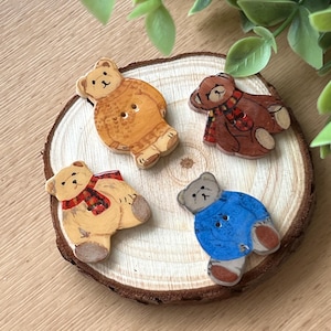 Può includere: Quattro bottoni di legno a forma di orso con dettagli dipinti. Gli orsi indossano maglioni e sciarpe di diversi colori. I bottoni sono disposti su una fetta di legno.