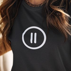 Può includere: Una donna indossa una maglietta grigio scuro con un simbolo di pausa bianco, un cappello da cowboy bianco e una cintura marrone. Sorride e sta in piedi accanto a un cavallo bianco. Lo sfondo è un campo e montagne.