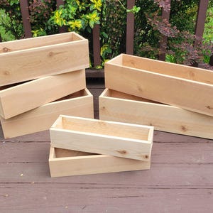 May include: Several rectangular wooden planters are stacked on a wooden deck. The planters are made of light-colored wood and have a simple, open design. The planters are empty and ready for planting flowers or herbs.