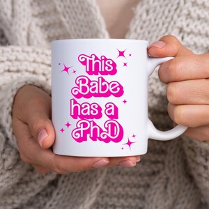 Pode incluir: Caneca de cerâmica branca com o texto "This Babe has a PhD" em rosa, juntamente com detalhes de estrelas rosa. A caneca é segurada por uma pessoa que usa um suéter de cor clara. A caneca tem uma alça.