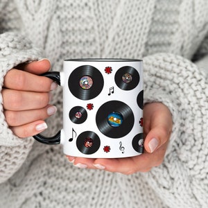 May include: A white ceramic mug with a black rim and handle. The mug is decorated with a pattern of black vinyl records and red four-leaf clover symbols. There are also musical notes printed on the mug.