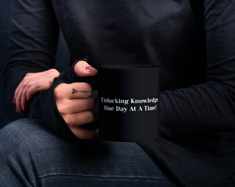 Puede incluir: Una taza de café negra con texto blanco que dice "Unlocking Knowledge One Day At A Time!" La taza está siendo sostenida por una persona que lleva una camisa negra de manga larga y pantalones vaqueros azules.