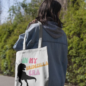Puede incluir: Una bolsa de lona blanca con una silueta negra de un dinosaurio con una corona y el texto "In My Bridezilla Era" en letras de colores.