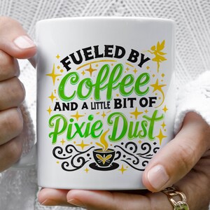 Puede incluir: Taza de cerámica blanca con un diseño en negro y verde que dice "Fueled by Coffee and a Little Bit of Pixie Dust". El diseño también incluye una pequeña estrella amarilla y un hada verde con alas.