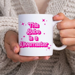 Könnte beinhalten: Weiße Keramiktasse mit rosa Schriftzug "This Babe is a Divemaster" mit rosa Sternen um den Text.